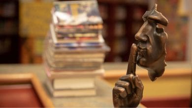 una maschera di rame che imita le fattezze di una faccia umana che indica di fare silezio, libri sullo sfondo