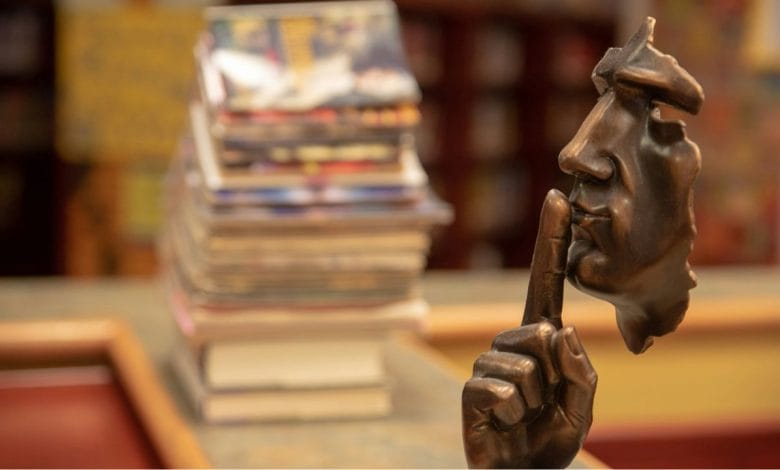 una maschera di rame che imita le fattezze di una faccia umana che indica di fare silezio, libri sullo sfondo