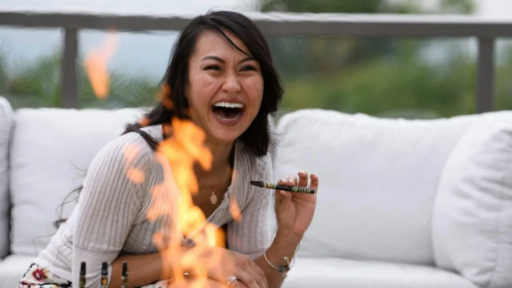 une femme asiatique riant de bon cœur devant un feu de camp tout en tenant une cigarette électronique