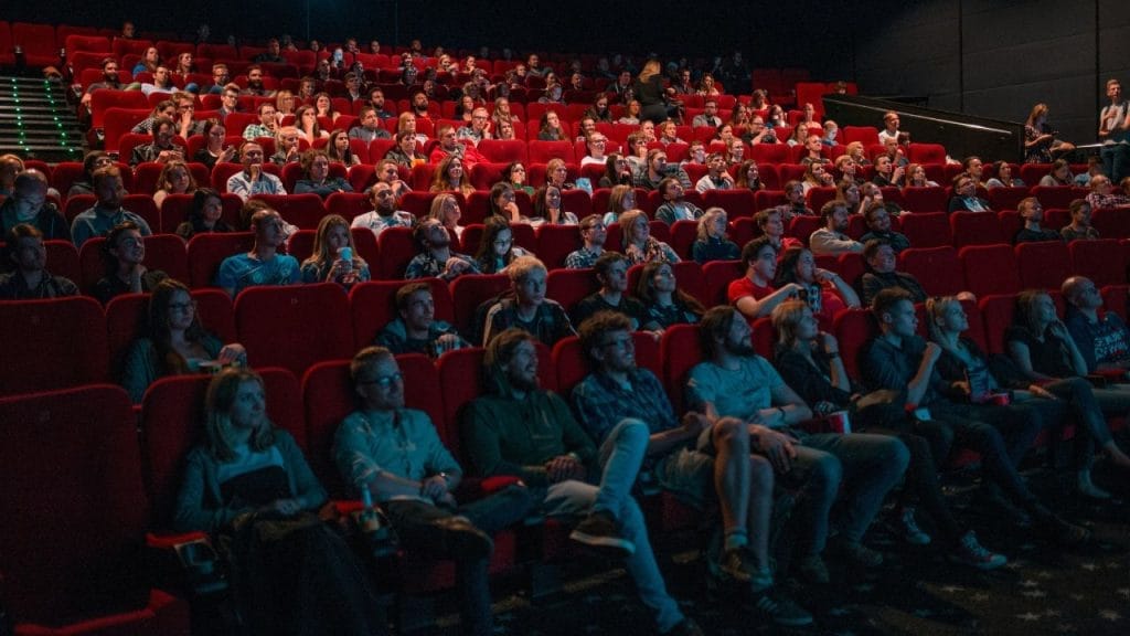 daug žmonių sėdinčių raudonose kino teatro kėdėse, žiūrintys rodomą filmą