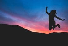 una donna che salta felice con in background un tramonto rosa sul cielo blu